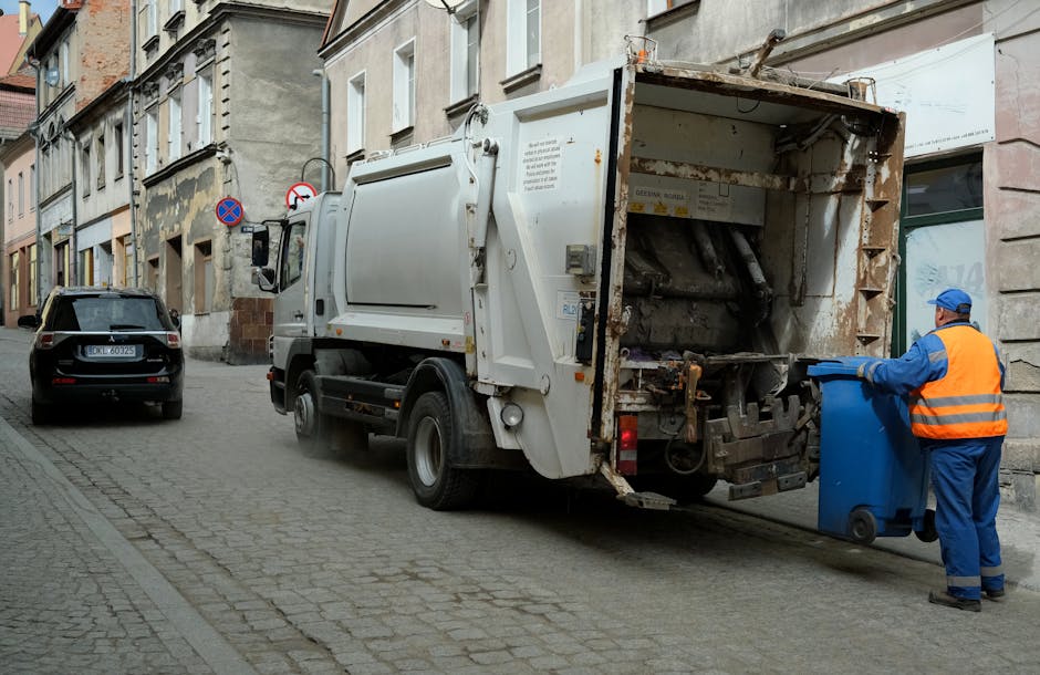 A street scene shows a commercial waste collection vehicle with an open rear compartment, positioned on a cobblestone road next to a row of residential buildings. A worker wearing a high-visibility orange vest and blue uniform is emptying a blue recycling bin into the truck’s compartment, which appears rusty and used. The surroundings include a black parked car, building facades with windows and doors, and street signage. The area is clean, with no visible debris on the road, and soft natural lighting illuminates the scene, emphasizing the urban environment and waste disposal process linked to bulky waste and mattress disposal services provided by Carpet Cleaners SW15 in SW15.