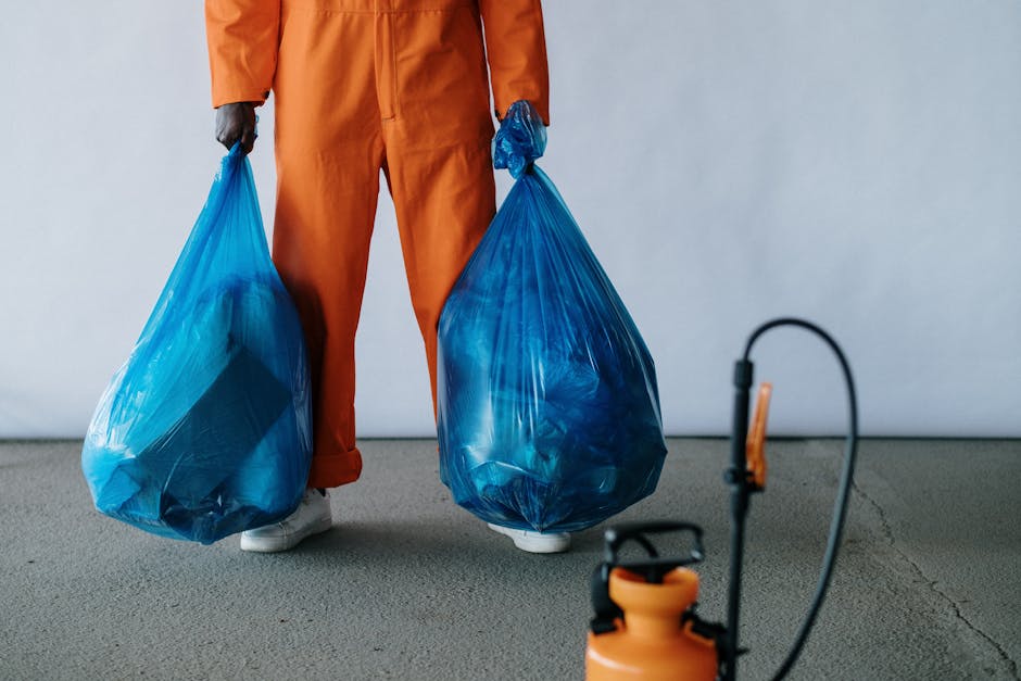 A person dressed in an orange jumpsuit holding two large blue plastic bags filled with bulky waste or debris, standing on a smooth concrete or vinyl floor next to a white wall. The individual is partially visible, with only their legs and hands shown. In the foreground, there is a small orange and black sprayer or cleaning device placed on the floor, suggesting a professional cleaning or waste disposal setting. The scene is well-lit with neutral lighting, emphasizing the cleanliness and organization typical of a commercial or domestic cleaning environment. Carpet Cleaners SW15 provides surface cleaning, deep cleaning, and sanitisation services for waste removal and mattress disposal in the SW15 area, as indicated by the page titled 'Bulky waste, mattress disposal and cleaning in SW15, SW15' on their website.