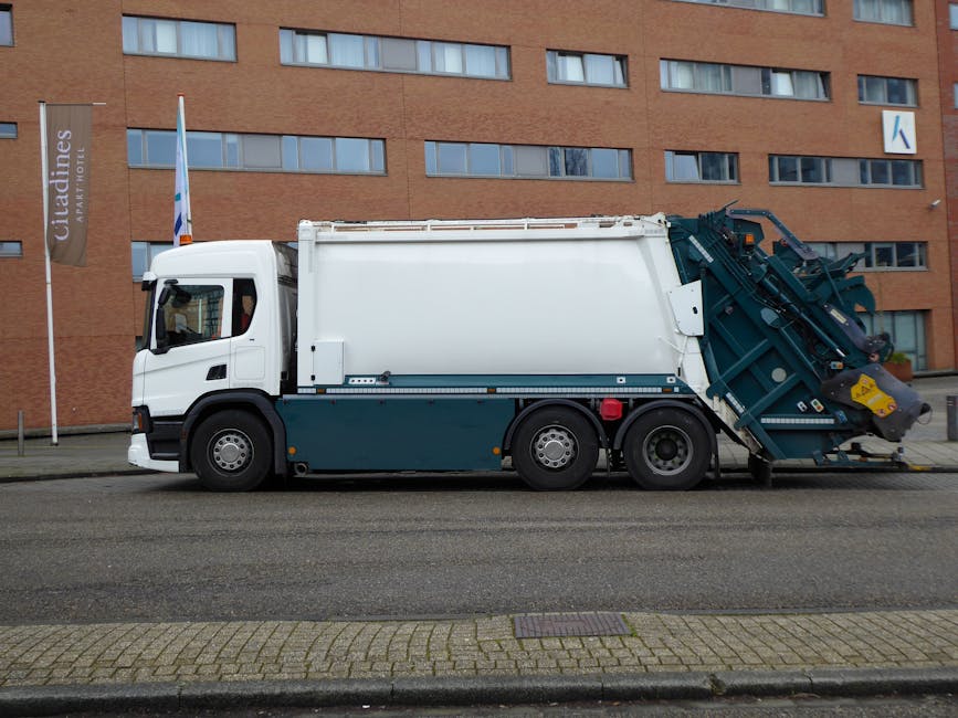 A street scene shows a commercial waste collection vehicle with an open rear compartment, positioned on a cobblestone road next to a row of residential buildings. A worker wearing a high-visibility orange vest and blue uniform is emptying a blue recycling bin into the truck’s compartment, which appears rusty and used. The surroundings include a black parked car, building facades with windows and doors, and street signage. The area is clean, with no visible debris on the road, and soft natural lighting illuminates the scene, emphasizing the urban environment and waste disposal process linked to bulky waste and mattress disposal services provided by Carpet Cleaners SW15 in SW15.