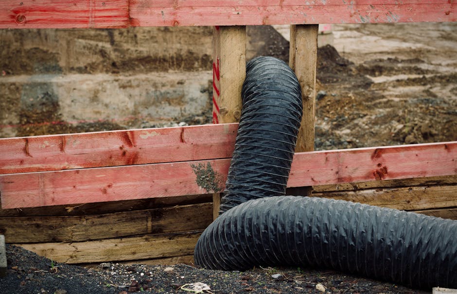 Close-up view of a black flexible duct pipe, used for ventilation or exhaust, emerging through a wooden frame with horizontal red-painted planks at a construction site. The background reveals exposed brick and soil, with natural lighting highlighting the industrial setting. The condition of the surfaces appears new and intact, with no visible dust or debris on the pipe or surrounding wood, indicating recent installation or maintenance. This image exemplifies surface cleaning and installation processes in a building or renovation context, relevant to professional cleaning and odour removal services offered by Carpet Cleaners SW15 near Putney Bridge, SW15, as part of their comprehensive cleaning and sanitisation solutions.