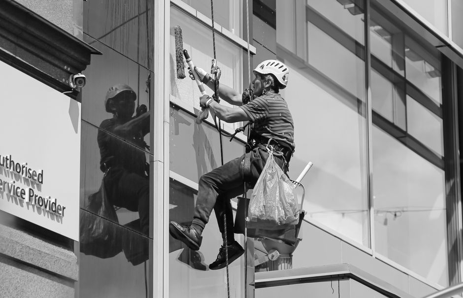 A professional window cleaner wearing a safety helmet and harness is cleaning the exterior glass façade of a modern commercial building in Putney, SW15. The worker is using a squeegee and a cleaning 