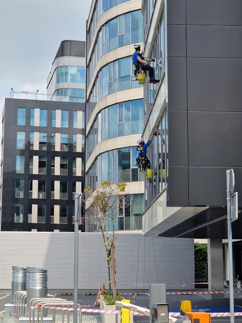 A professional window cleaner wearing a safety helmet and harness is cleaning the exterior glass façade of a modern commercial building in Putney, SW15. The worker is using a squeegee and a cleaning 