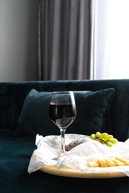 A close-up of a dark teal sofa in a residential living room, with a matching cushion visible behind a clear glass of red wine placed on a white paper napkin. The sofa is positioned against a wall with floor-length grey curtains and a white window frame that allows natural light to illuminate the scene. Adjacent to the wine glass, there is a beige serving tray holding a bunch of green grapes and slices of cheese, indicating a setting for relaxation or entertaining. The overall environment appears clean and well-maintained, suitable for domestic surface cleaning and deep cleaning services provided by Carpet Cleaners SW15, in connection with quick fix solutions for stains on Putney sofas, SW15.