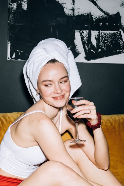 A young woman with a white towel wrapped around her head, sitting on a mustard yellow sofa in a living room. She is wearing a white tank top and has a red scrunchie on her wrist. She holds a glass of red wine in her right hand and is smiling with a slight smirk, looking to her left. Behind her, there is a dark wall with a large black and white abstract painting. The surface appears clean and well-maintained, with soft lighting highlighting her face and upper body. The scene suggests a relaxed, domestic setting, possibly after a deep cleaning session or household refresh, consistent with the themes of surface cleaning and maintenance associated with the page's focus on quick fixes for stains like red wine on sofas, as covered by Carpet Cleaners SW15.