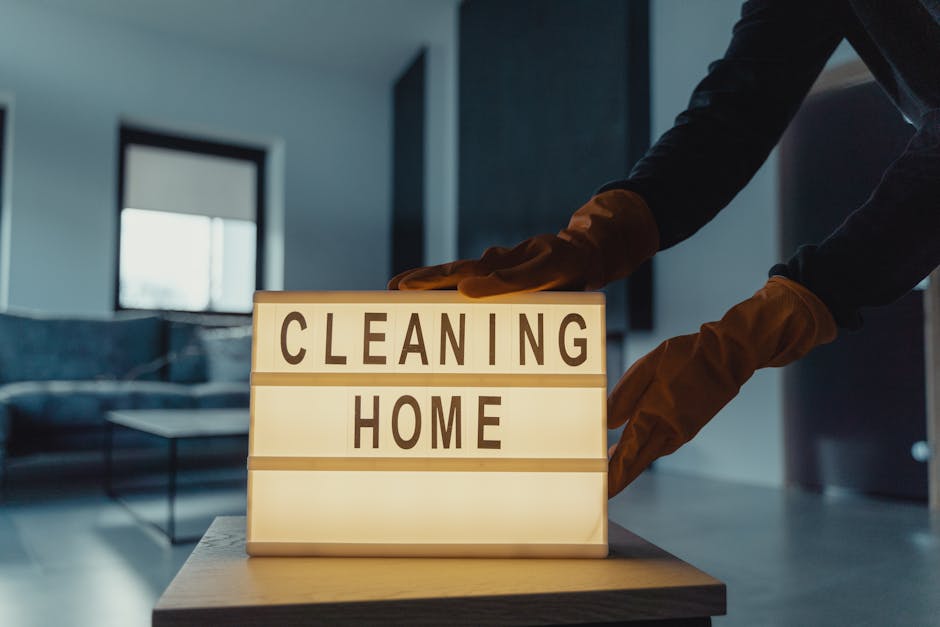 A person wearing brown cleaning gloves is adjusting a lit-up sign on a wooden table that displays the words 'CLEANING HOME'. The background shows a modern living room with a large window letting in natural light, a dark sofa, a coffee table, and a contemporary interior design. The surface of the sign is smooth, and the room appears clean and tidy, reflecting professional domestic cleaning services by Carpet Cleaners SW15, aligned with their deep-cleaning and sanitisation expertise highlighted in the SW15 upper Richmond Road homes guide.
