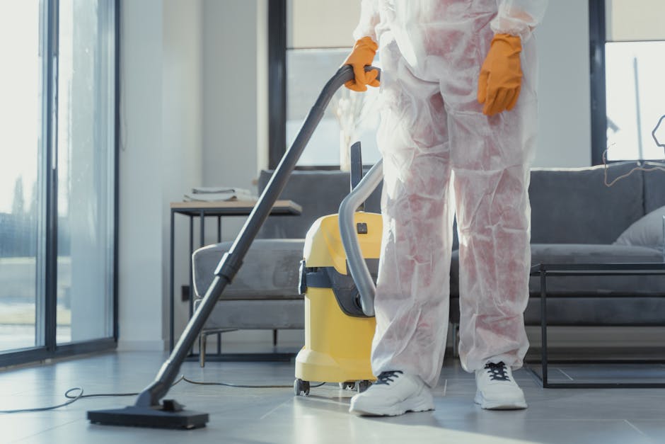 A professional cleaner from Carpet Cleaners SW15 is engaged in surface cleaning within a modern living room. The person, dressed in a white protective suit and yellow gloves, operates a yellow and gray vacuum cleaner on a tiled floor, showcasing a deep-cleaning process. The room features a glass sliding door, allowing natural light to illuminate the space, alongside a gray fabric sofa with a cushion and a small side table with a few books. The tiled surface is clean and wet, indicating recent scrubbing or sanitisation, and the environment emphasizes hygiene and meticulous domestic cleaning procedures. The image highlights the use of specialized cleaning tools in maintaining a spotless and hygienic home environment, aligning with surface and deep cleaning practices promoted by Carpet Cleaners SW15 on the webpage for Upper Richmond Road homes.