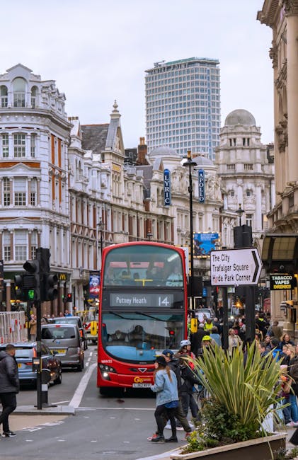 A busy street scene in Wandsworth, featuring a red double-decker bus labeled 'Putney Heath 14' approaching a pedestrian crossing. The area is surrounded by historic white buildings with ornate facades and large windows, alongside modern high-rise structures. Pedestrians are crossing the street, some waiting on the sidewalk near potted plants and small trees. The sky is overcast, casting diffused light over the scene. Visible storefronts include a tabacco shop and various shops, with street signs pointing toward Trafalgar Square and Hyde Park Corner, indicating central London locations. The traffic includes cars, taxis, and cyclists, illustrating a vibrant urban environment. This image exemplifies the typical surface cleaning and maintenance of busy city streets, as managed by local authorities and cleaning services like Carpet Cleaners SW15, who specialize in thorough cleaning, sanitisation, and hygiene maintenance of urban surfaces.