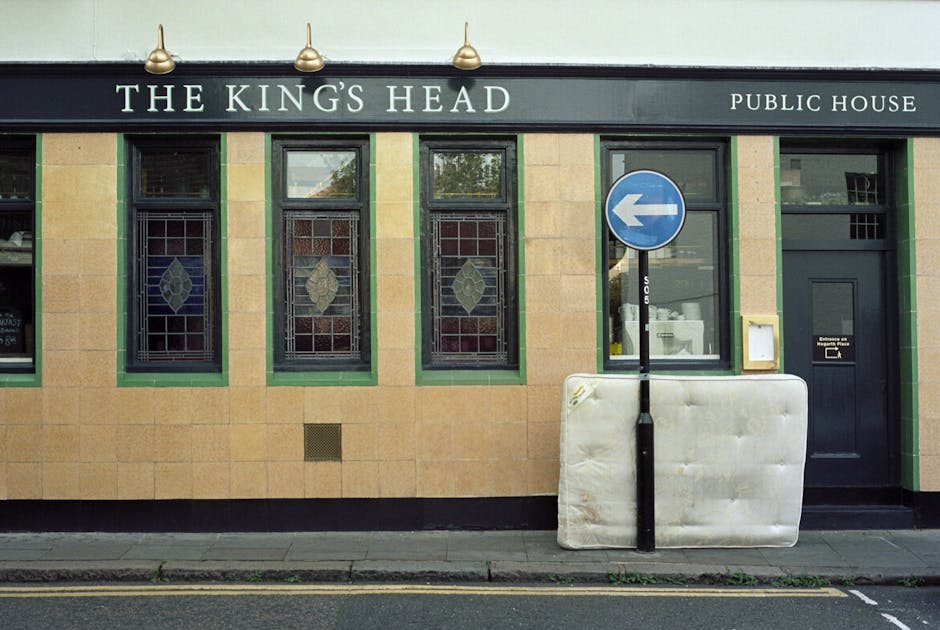 The exterior of The King's Head public house features a beige brick facade with four stained glass windows framed in green, a black door on the right, and a blue traffic sign indicating a left turn positioned near a large, white, foam-filled barrier leaning against a black pole. The signage above the windows displays the pub's name in white letters on a dark background. The building is illuminated by three small, gold-colored exterior lights. The sidewalk in front appears clean, with subtle cracks and patches of grass between paving stones. The scene is well-lit with natural daylight, highlighting the cleanliness and neat appearance of the surroundings, reflecting a well-maintained street environment, relevant to surface cleaning and maintenance for commercial properties like pubs and public houses.